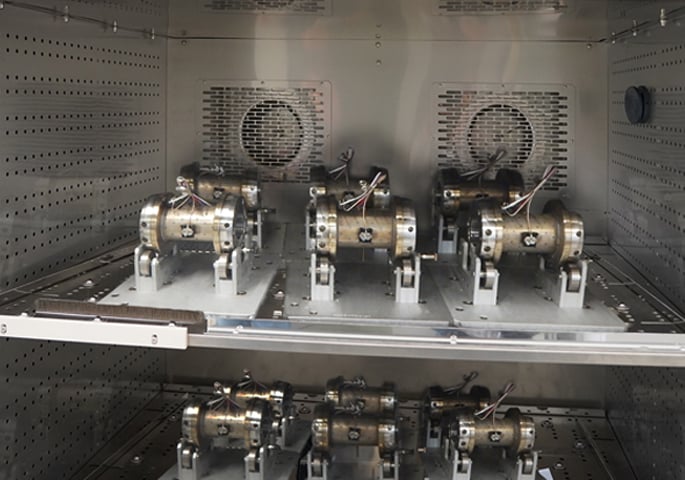 Metal workpieces on holders in a climate test chamber with fans on the rear wall.