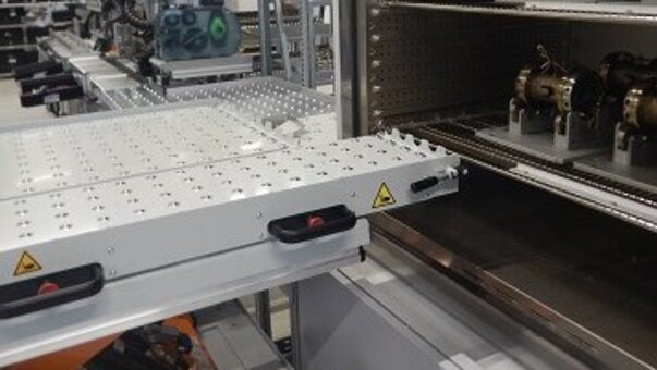 Industrielle Produktionsanlage mit offenem Prüfschrank im Vordergrund, in dem sich Werkstücke befinden. Eine Hebebühne steht vor dem Prüfschrank.