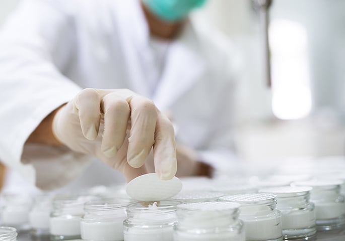 Proben kosmetischer Creme in einem Labor. Im Hintergrund ein Labormitarbeiter in mit Handschuhen und Mundschutz.