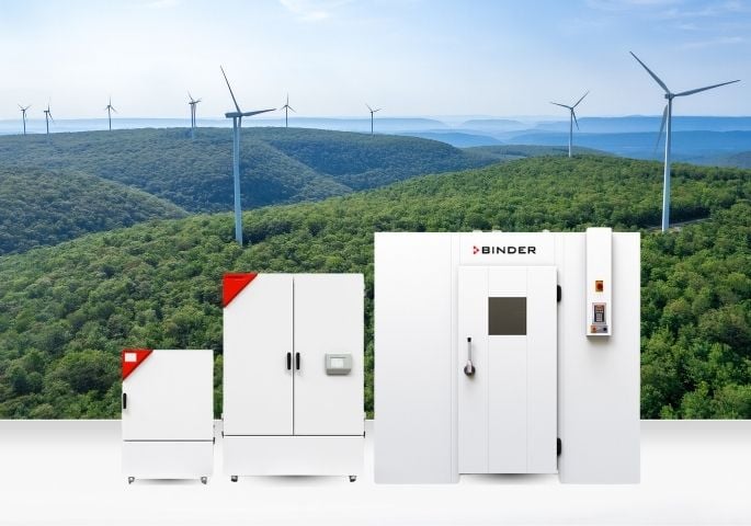 BINDER climate chambers in front of a landscape with wooded hills and wind turbines in the background.